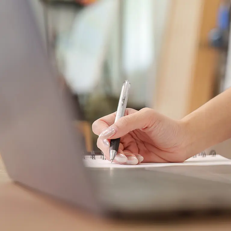 hand writing with a pen on a notebook next to an open laptop