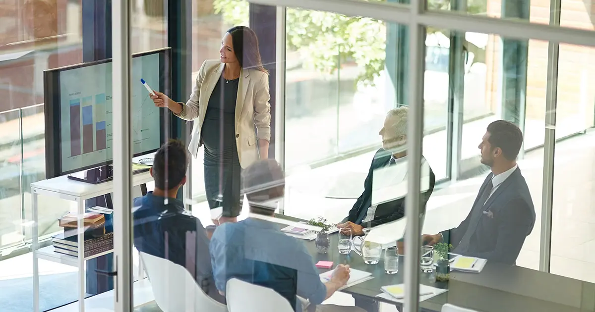 people in a board room listening to someone present at the front pointing to a screen