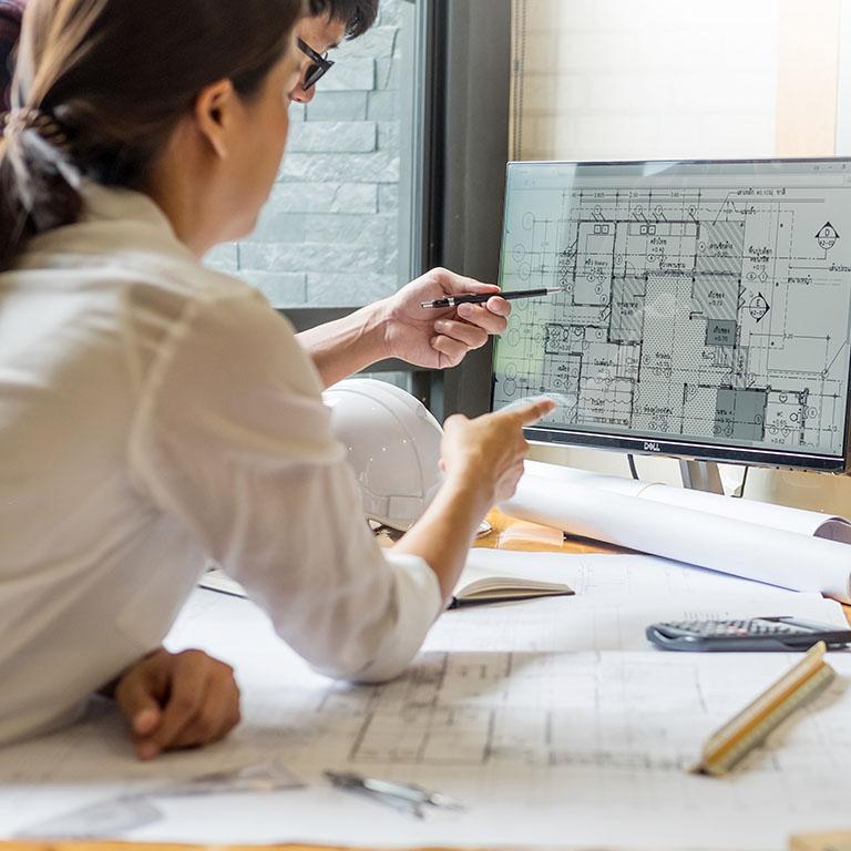 two people pointing to a monitor with plans on a desk